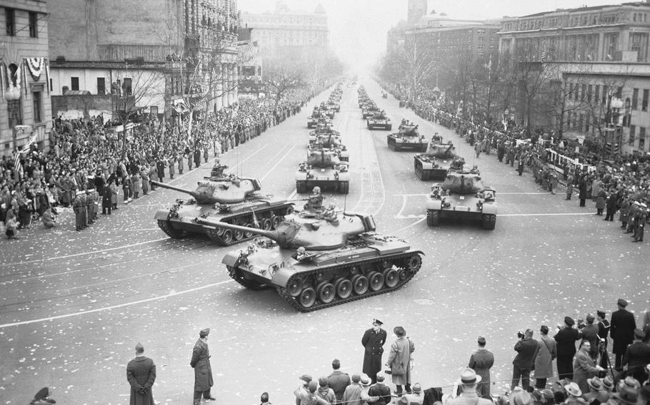 A black-and-white, 1950s-era image of Army tanks traveling down a city street with crowds of spectators lining either side.