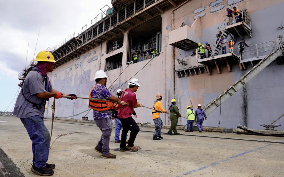 Several men in hard hats on a pier pull a line as they work on a ship.