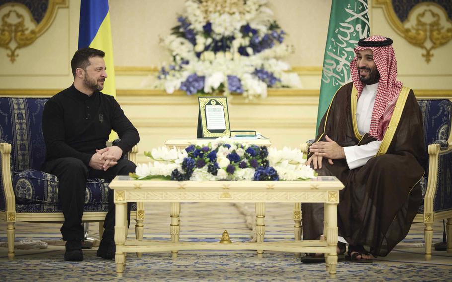 Ukrainian President Volodymyr Zelenskyy and Saudi Arabian Crown Prince Mohammed bin Salman sit in blue-cushioned chairs on opposite sides of a white table with a flowered wreath on top.