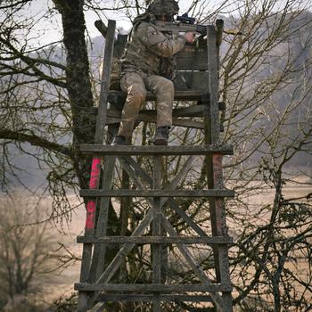 A U.K. soldier points a rifle from on top of a wooden lookout.