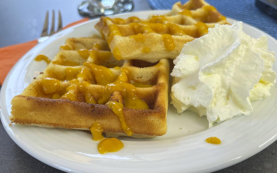 A white plate with golden waffles drizzled in an orange colored sauce, accompanied by a dollop of whipped cream. 
