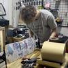 A man hunches over a table to make repairs on a skateboard.