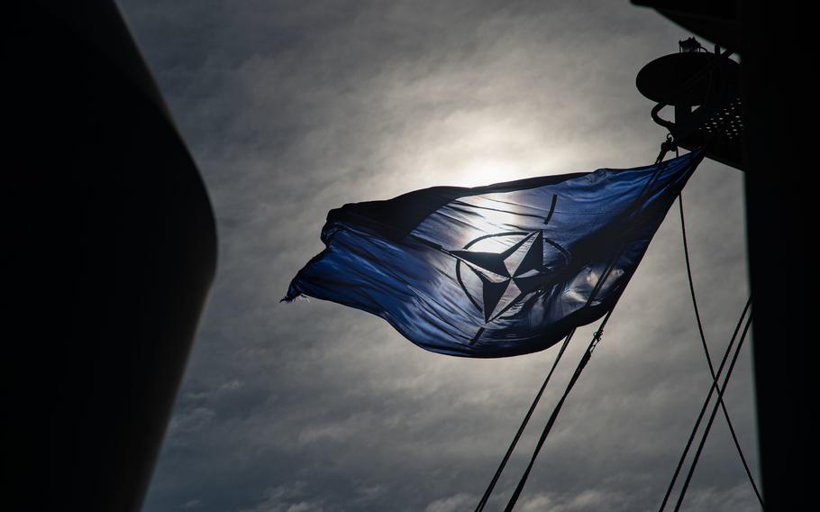 The NATO flag flies against a cloudy sky.