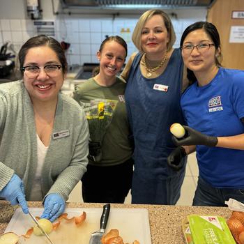 Group of four women standing together smiling for camera. The woman on the far left has blue medical gloves on and is slicing an onion on a white chopping board.