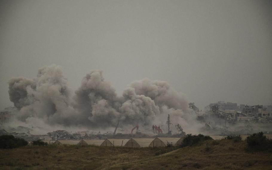 Smoke billows over a landscape of destroyed buildings, with some tents visible in the foreground.
