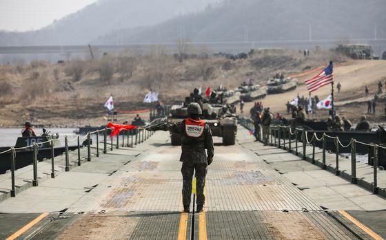 A South Korean soldier in camouflage uniform stands on a pontoon bridge holding red safety flags, directing military vehicles including tanks and armored personnel carriers crossing the bridge. There are American and South Korean flags visible in the background. Mountains and hazy terrain can be seen in the distance, and the soldier is wearing an orange safety vest.