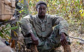 A soldier in a camouflage uniform sits on the ground in a wooded area with his arms resting on his knees.