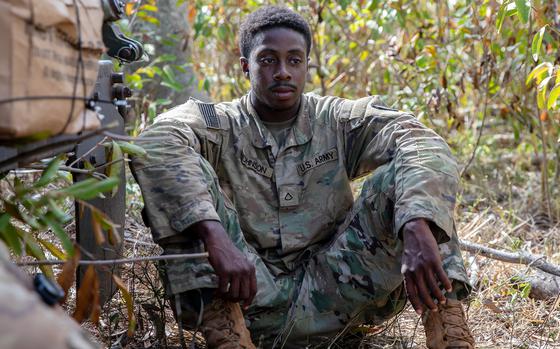 A soldier in a camouflage uniform sits on the ground in a wooded area with his arms resting on his knees.
