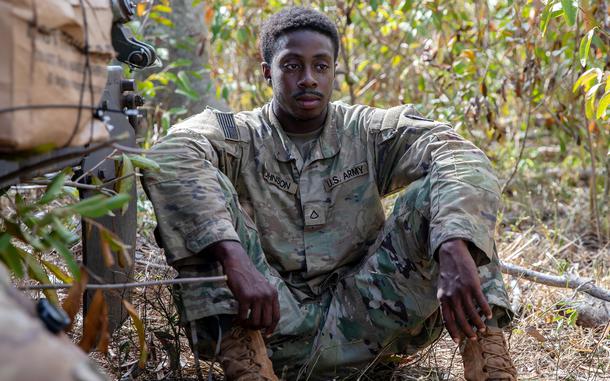 A soldier in a camouflage uniform sits on the ground in a wooded area with his arms resting on his knees.
