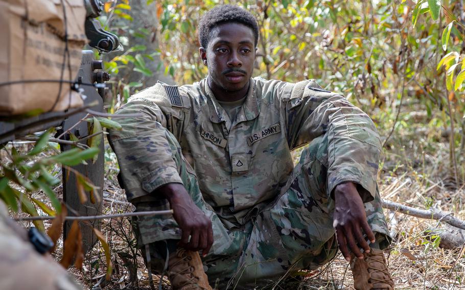 A soldier in a camouflage uniform sits on the ground in a wooded area with his arms resting on his knees.