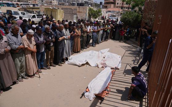 A group of Palestinian mourners stand in a line with their heads bowed in prayer in front of a body on the ground wrapped in a white shroud.