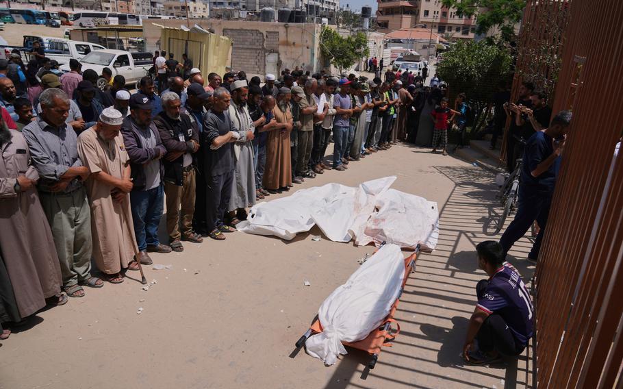 A group of Palestinian mourners stand in a line with their heads bowed in prayer in front of a body on the ground wrapped in a white shroud.
