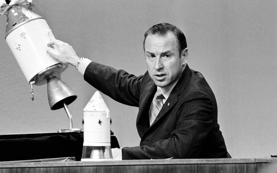 A black-and-white 1970s-era image of a man in a dark suit seated at a desk and pointing to a spot on a model of a space craft.