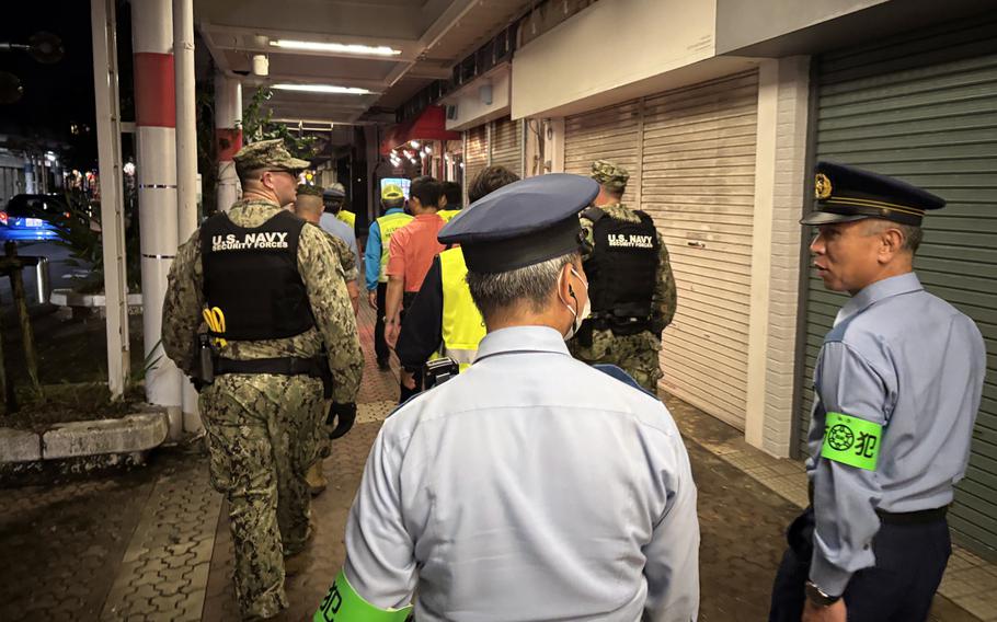 Military police and Japanese police walk together past shuttered storefronts at night.