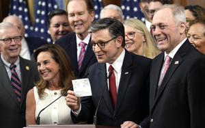 House Speaker Mike Johnson stands at a podium holding up a white card or paper with numbers ‘218 214’ written on it, surrounded by smiling colleagues in formal attire. 