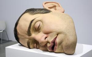 A large, life-like sculpture of a man’s head lying on its cheek rests on a white platform in a museum.
