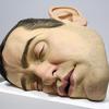 A large, life-like sculpture of a man’s head lying on its cheek rests on a white platform in a museum.