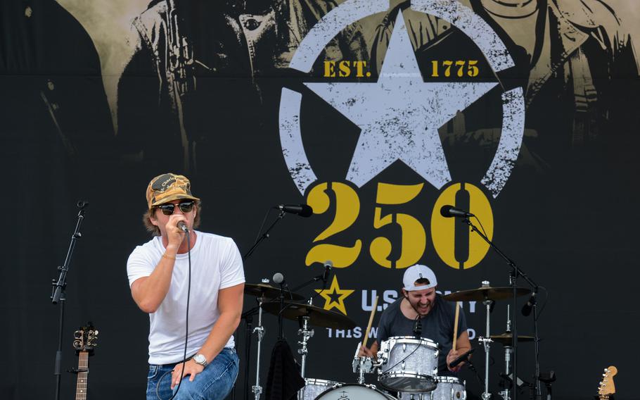 A singer holds a microphone and leans forward on stage with a drummer playing behind him.