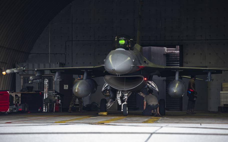 Military aircraft maintenance in a hangar showing a fighter jet undergoing service. Ground crew personnel are working around the aircraft, which is positioned on the hangar floor with external fuel tanks visible. 