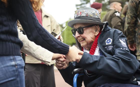 An elderly military veteran in a wheelchair wearing sunglasses and a military cap adorned with multiple service pins shakes hands with someone in a navy blue suit jacket. The veteran is wearing a navy blue jacket with military patches and a red, white and blue lei around his neck. Other veterans and attendees can be seen in the blurred background.