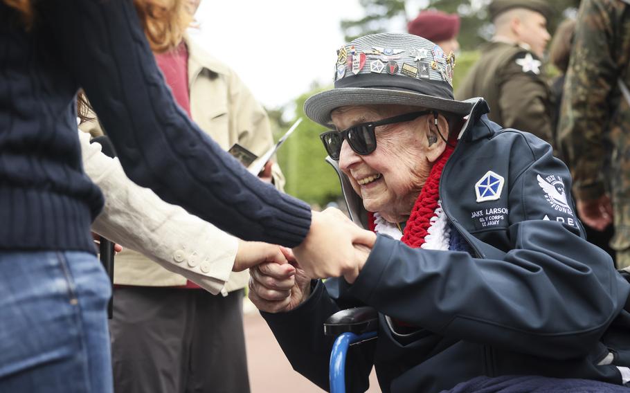 An elderly military veteran in a wheelchair wearing sunglasses and a military cap adorned with multiple service pins shakes hands with someone in a navy blue suit jacket. The veteran is wearing a navy blue jacket with military patches and a red, white and blue lei around his neck. Other veterans and attendees can be seen in the blurred background.