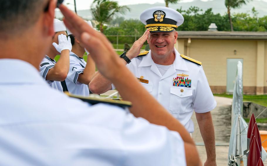A smiling Navy officer in dress white uniform with captain’s hat and gold braiding returns a salute to sailors in white uniforms. The scene takes place outdoors with palm trees and mountains visible in the background.
