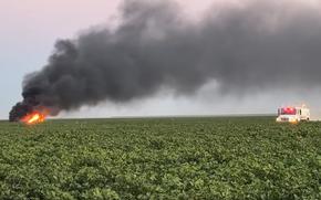 A rescue vehicle cruises through the cotton field toward the site of a jet crash. Black smoke rises from the flaming jet.