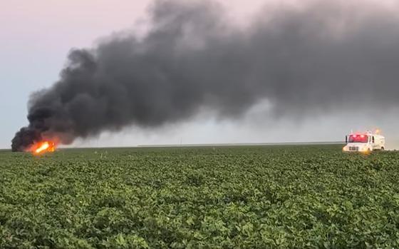 A rescue vehicle cruises through the cotton field toward the site of a jet crash. Black smoke rises from the flaming jet.