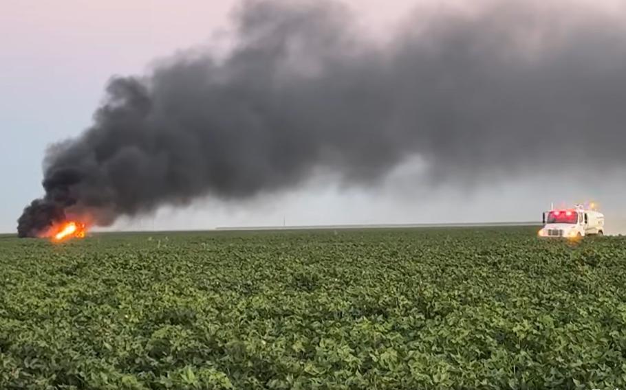 A rescue vehicle cruises through the cotton field toward the site of a jet crash. Black smoke rises from the flaming jet.