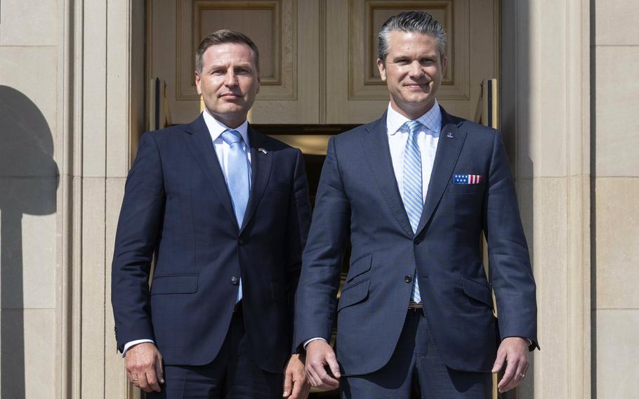 Two men in dark business suits and blue ties stand side by side in front of an building entrance. 
