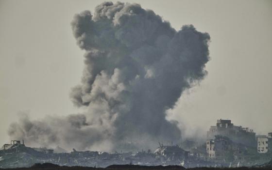 A large plume of smoke, seen from a distance, rises into the sky above a city skyline of damaged and destroyed buildings.