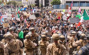 Troops in camouflage uniforms and helmets holding riot shields stand in front of a group of protesters in a plaza outside an office building.