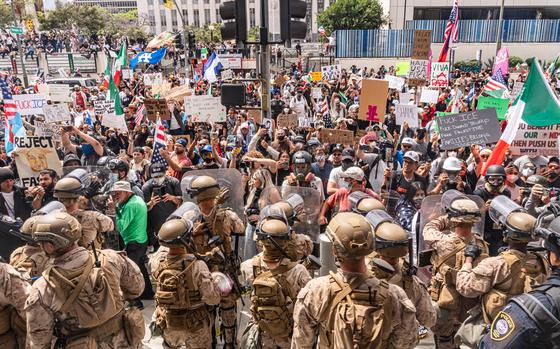Troops in camouflage uniforms and helmets holding riot shields stand in front of a group of protesters in a plaza outside an office building.
