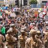 Troops in camouflage uniforms and helmets holding riot shields stand in front of a group of protesters in a plaza outside an office building.