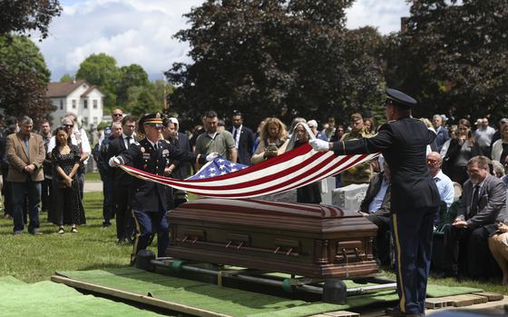 Two service members in uniform fold a flag above a casket at a cemetery.