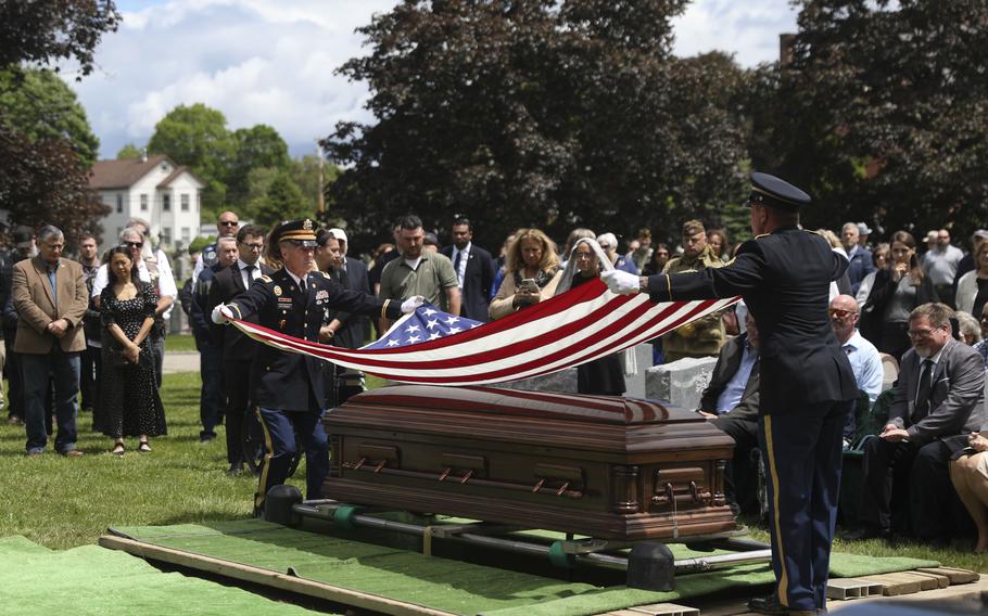 Two service members in uniform fold a flag above a casket at a cemetery.