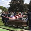 Two service members in uniform fold a flag above a casket at a cemetery.
