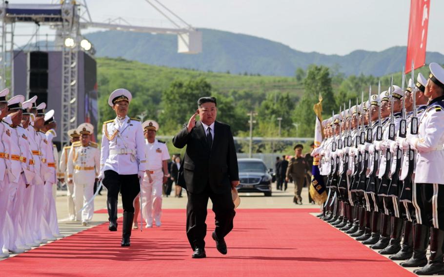 Kim Jong Un walks down an outdoor red carpet alongside a member of the North Korean navy as other military members lined up along the sides of the carpet watch.