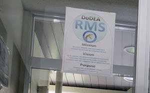 A poster inside a building displays the DoDEA RMS (Department of Defense Education Activity - Ryukyu Middle School) mission, vision, and purpose statements. The poster is mounted on a glass surface in a hallway or office area.