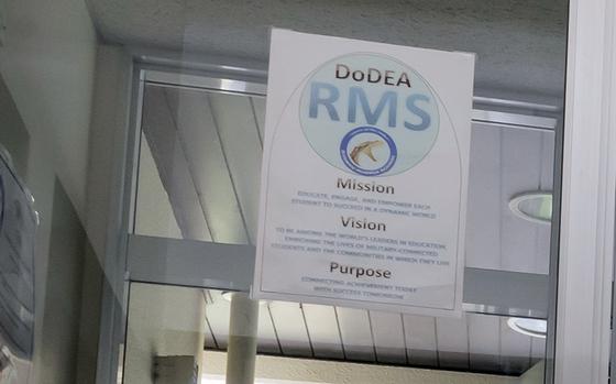 A poster inside a building displays the DoDEA RMS (Department of Defense Education Activity - Ryukyu Middle School) mission, vision, and purpose statements. The poster is mounted on a glass surface in a hallway or office area.