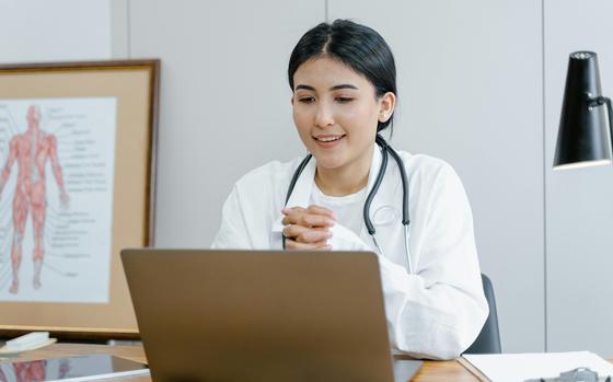A doctor in a white coat sits at a desk in front of their laptop.