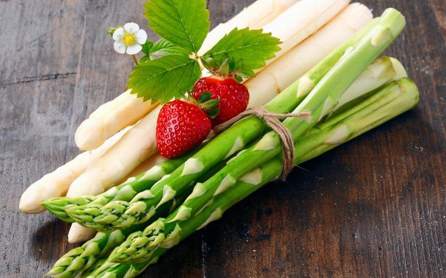 Bundles of green and white asparagus lie on a wooden surface with strawberries.