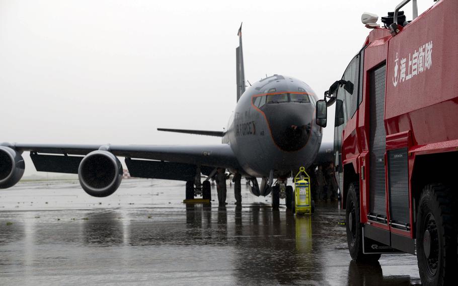 An aircraft sits on a wet airport tarmac with a red fire truck positioned nearby. The tarmac surface is wet, reflecting the overcast sky conditions. Japanese text is visible on the side of the fire truck.