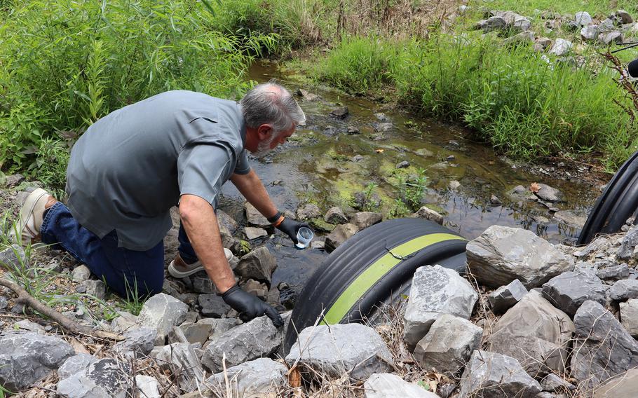 A man wearing a gray shirt and black gloves kneels down on rocks next to a creek and uses a small plastic container to collect a water sample.