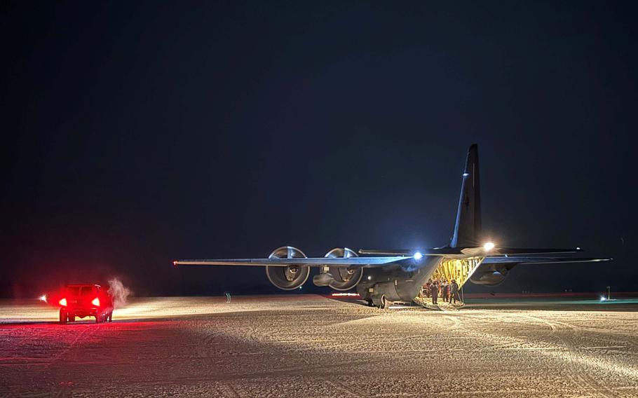 A military fighter jet sits on a runway at night with emergency vehicles nearby, their red lights illuminating part of the scene. 