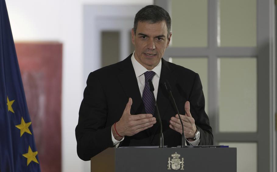 A man in a dark suit gestures with his hands as he stands at a podium and speaks into a microphone.