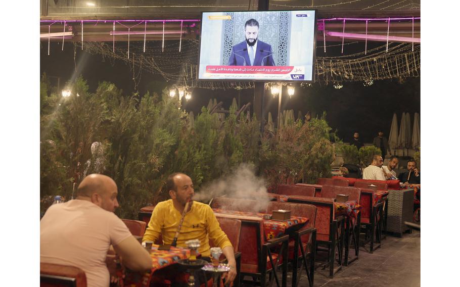 Syrians smoking hookah at a cafe, with a mounted TV above them showing the country’s president giving a speech.