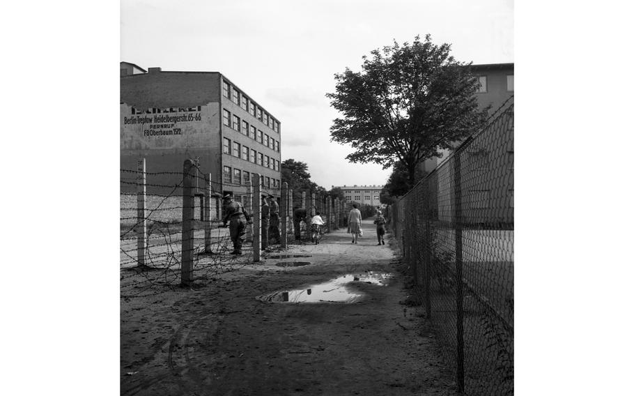 A West Berlin woman and her children walk along the border as uniformed East Germans roll out barbed wire.