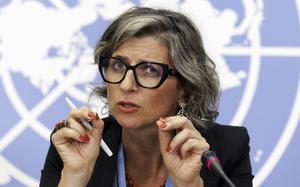 A close-up view of a woman in black-rimmed glasses gesturing with her hands while speaking into a microphone in front of a blue wall with the white logo of the United Nations.
