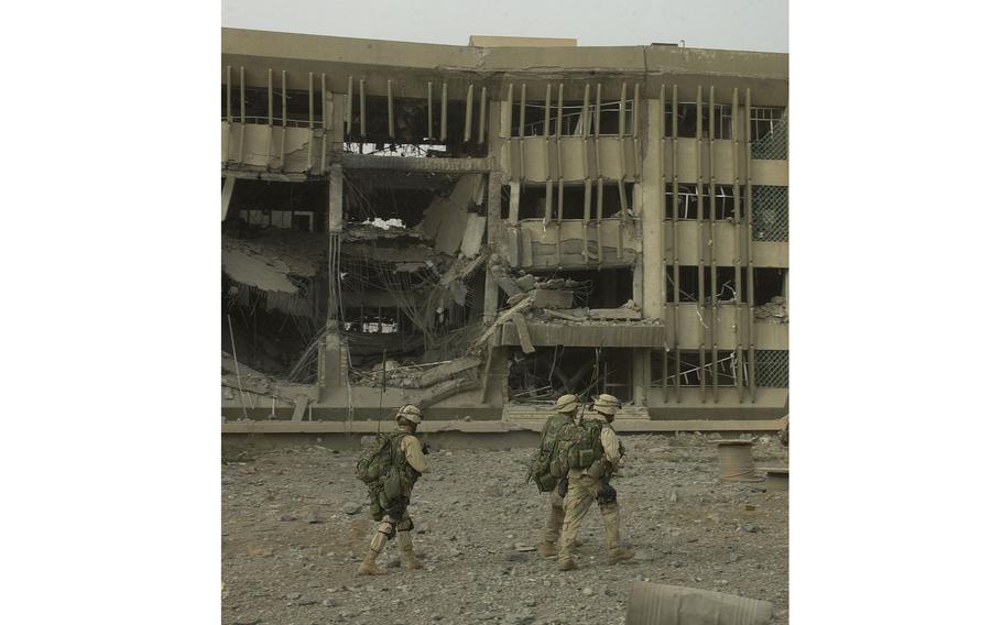 Marines in combat gear and holding weapons walk through a patch of sand and rocks in front of a heavily damaged building.
